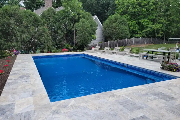 Rectangular in-ground swimming pool surrounded by light gray stone tiles, with lounge chairs and a picnic table on one side.