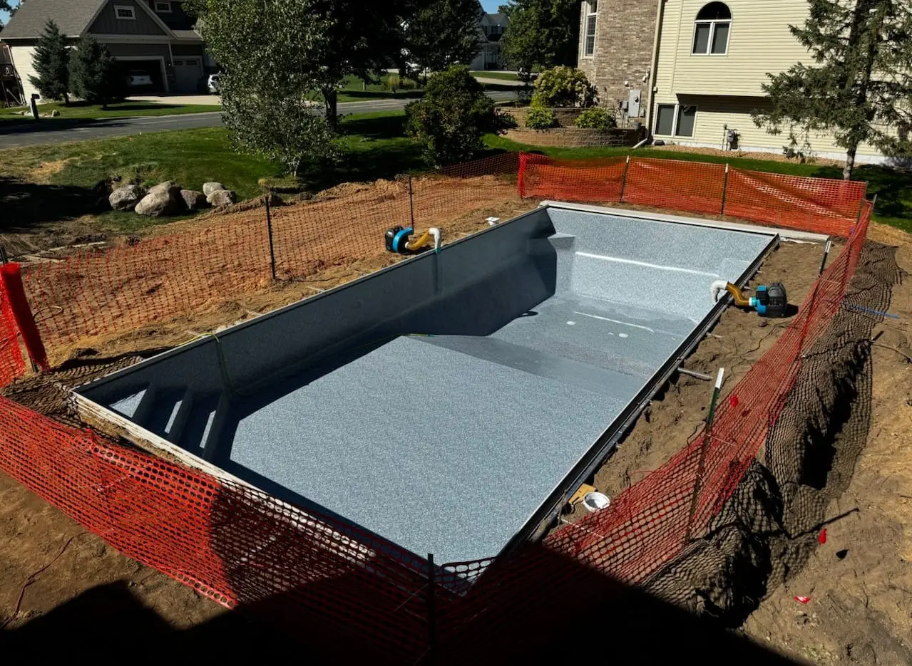 In-ground swimming pool installation underway in a Stillwater, MN backyard. The pool structure is fully set with a light blue patterned liner in place, and the surrounding dirt is exposed. Orange construction fencing encircles the site for safety.