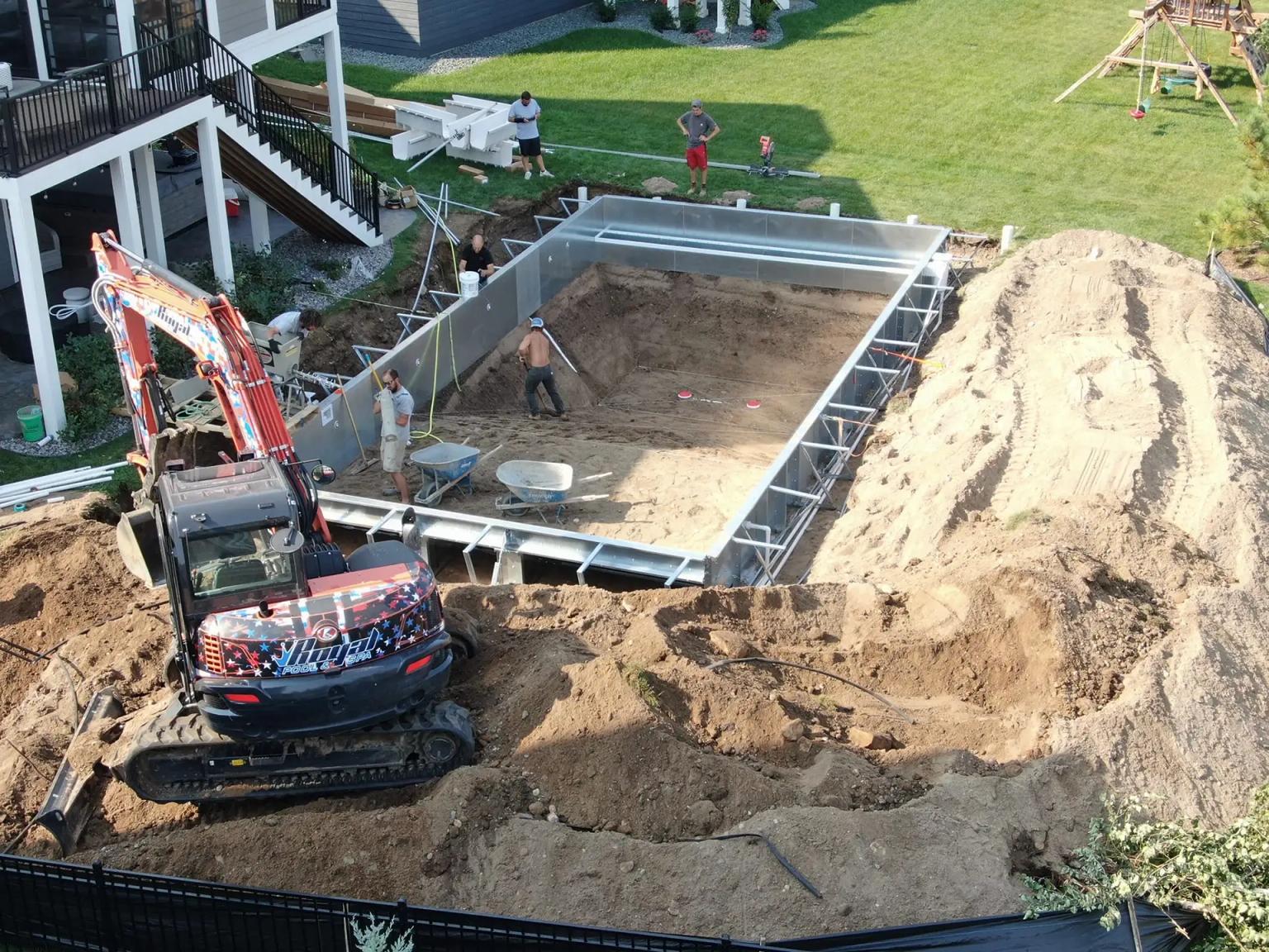 A residential backyard pool construction in White Bear Lake, MN showing workers assembling a rectangular in-ground pool. A Royal Pool and Spa logoed excavator is parked nearby, and the surrounding area is filled with dirt piles and construction materials.