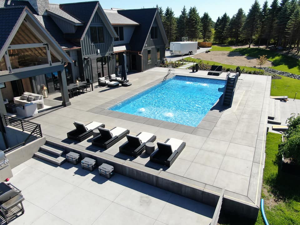 A luxurious modern backyard features a large rectangular swimming pool with clear blue water, surrounded by an expansive light gray concrete patio.