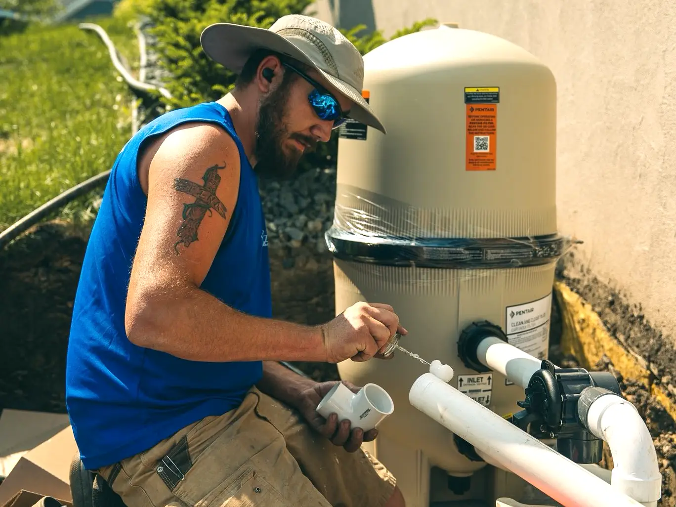 A man wearing a wide-brimmed hat, sunglasses, and a blue sleeveless shirt works on PVC piping next to a Pentair pool filtration system.