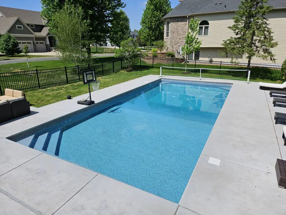 Backyard in-ground swimming pool surrounded by a smooth concrete deck, with patio furniture, a basketball hoop, and lounge chairs nearby. The pool is clean and clear, located in a residential neighborhood in Woodbury, MN.