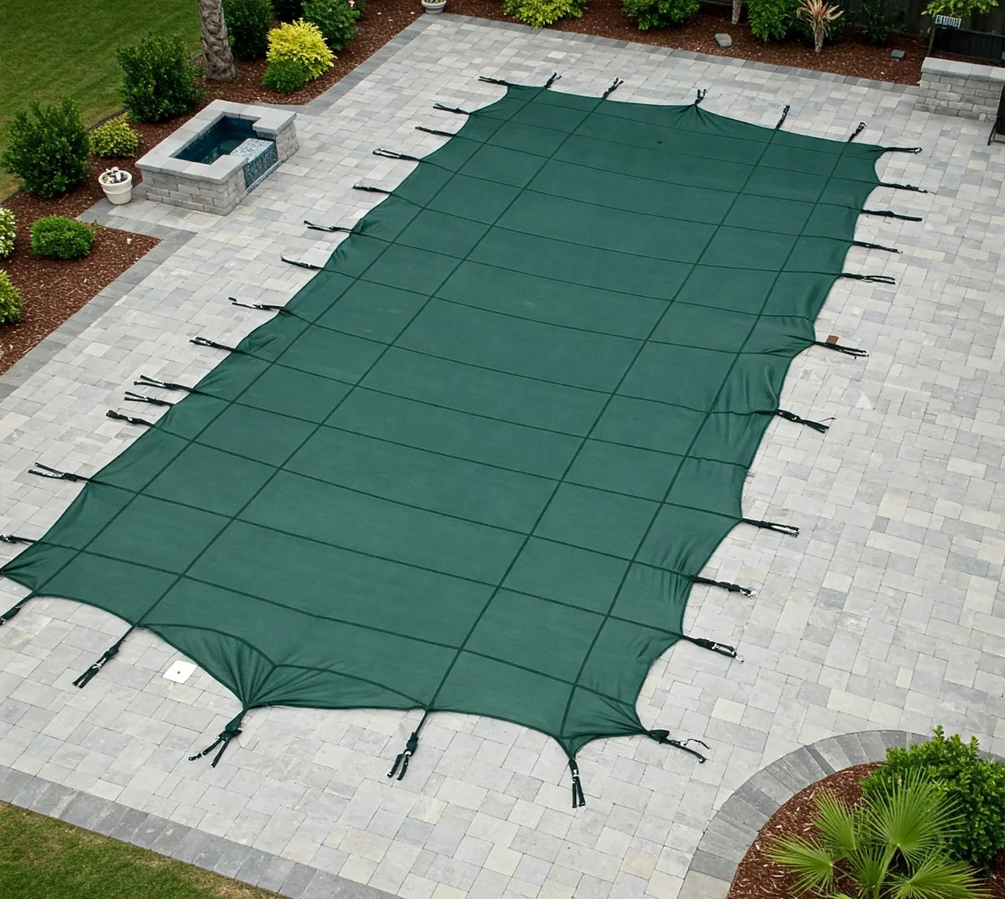 A rectangular inground swimming pool is covered with a taut green mesh safety cover, secured to the surrounding light gray stone paver patio with evenly spaced black straps and anchors.