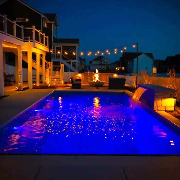 A backyard swimming pool at night is illuminated with vibrant blue underwater lighting, creating a striking glow. A waterfall feature flows into the pool from a stone structure on the right side.
