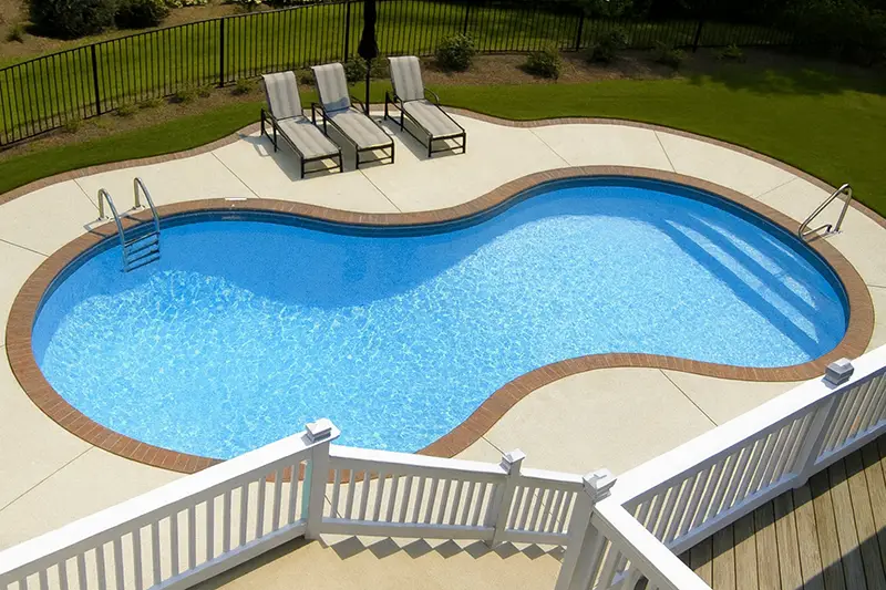 A bright aerial view of a figure-eight-shaped swimming pool with sparkling blue water, surrounded by a beige concrete deck edged with reddish-brown bricks.