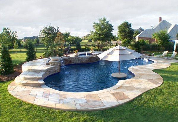 A freeform inground swimming pool with deep blue water features a natural, curving shape surrounded by a stone-tiled deck.
