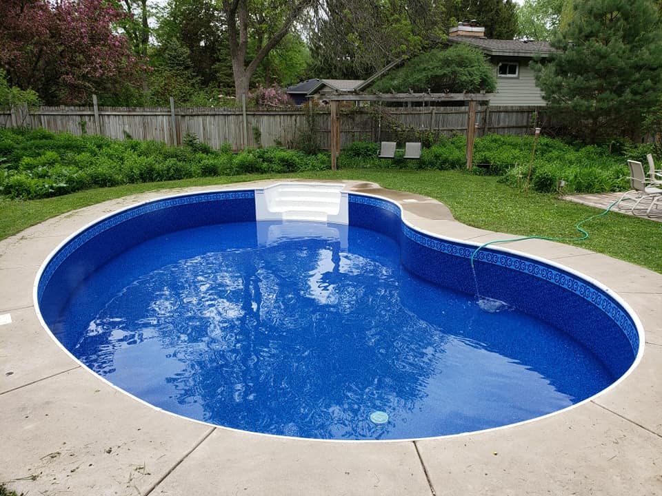 A kidney-shaped inground swimming pool with bright blue water and a decorative tile border sits in a backyard surrounded by greenery.