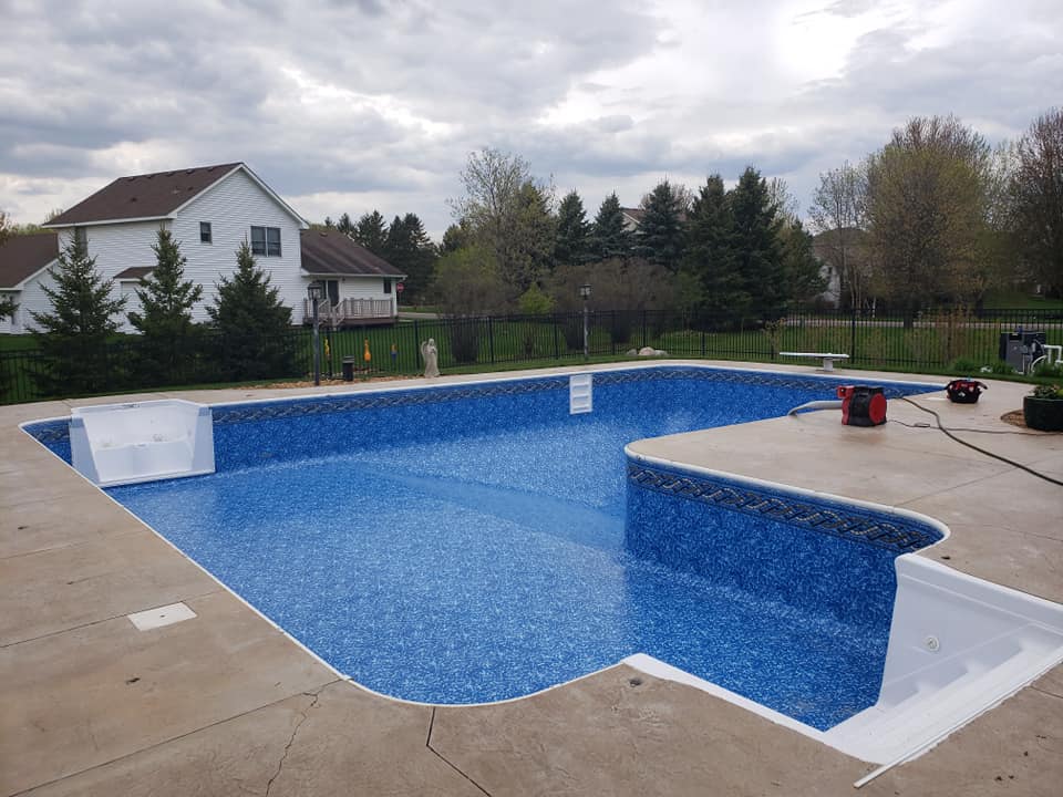 An empty L-shaped inground swimming pool in Centerville, MN with a blue patterned liner and white entry steps, sits in a backyard surrounded by a concrete deck.
