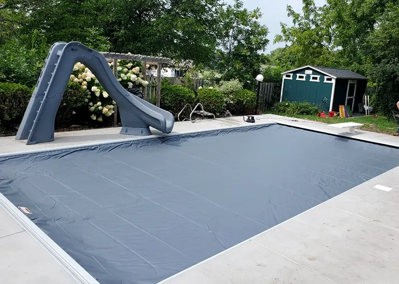 A rectangular inground swimming pool is covered with a smooth, gray automatic safety cover, surrounded by a light gray concrete deck.