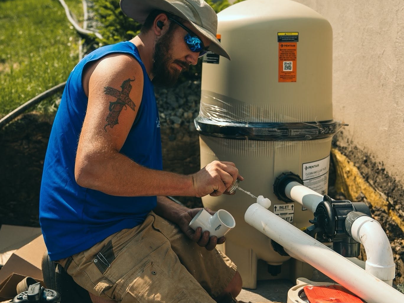 Swimming pool technician in a sun hat and safety glasses applies sealant to white PVC pipes beside a Pentair pool filtration unit.