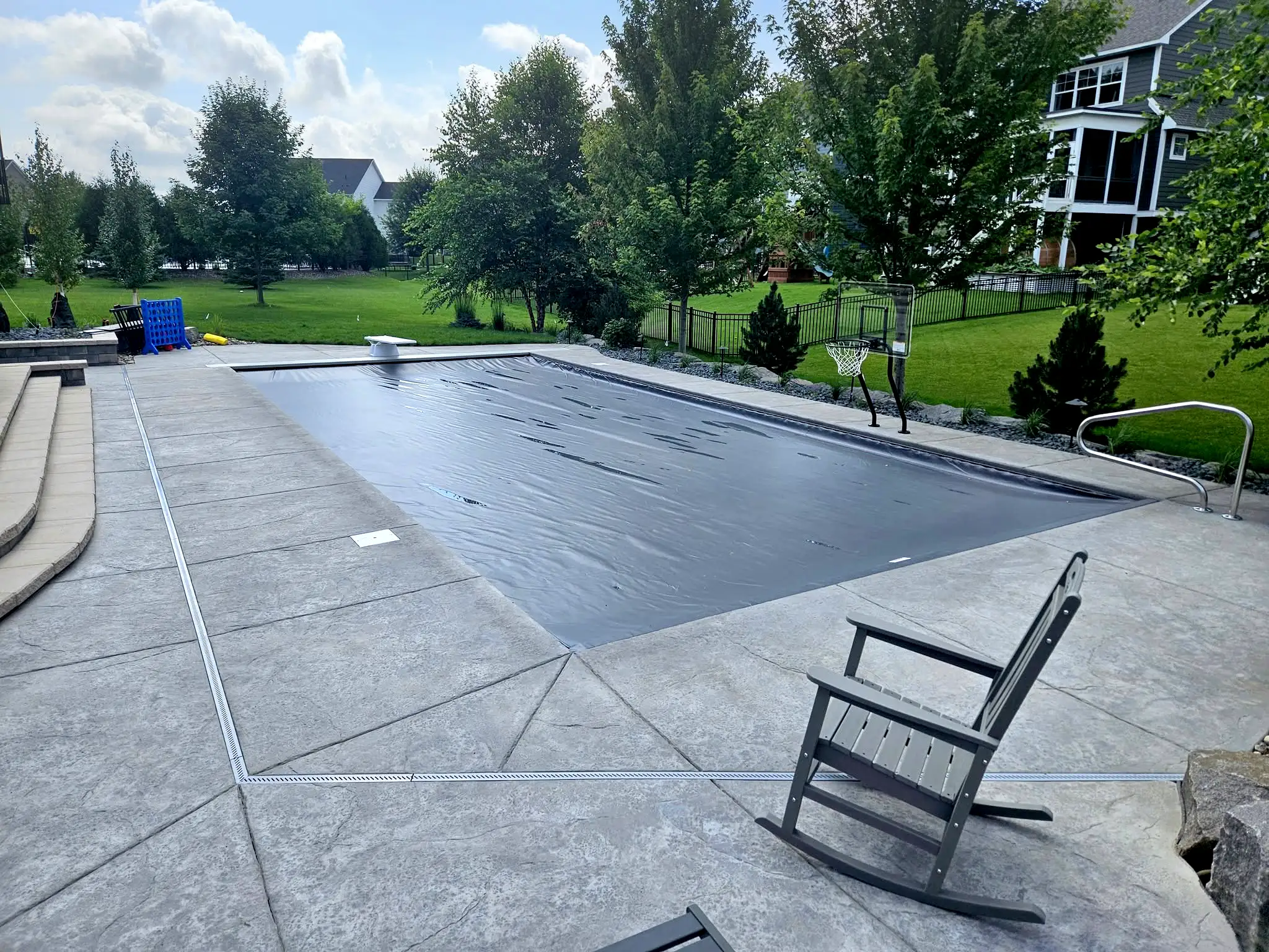 A modern backyard pool is covered with a dark gray safety cover, surrounded by stamped concrete decking and neatly landscaped greenery